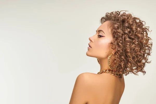 Jewelry and beauty. Portrait of a beautiful tanned girl with fine curls and elegant makeup with arrows posing in gold jewelry. White background with copy space.