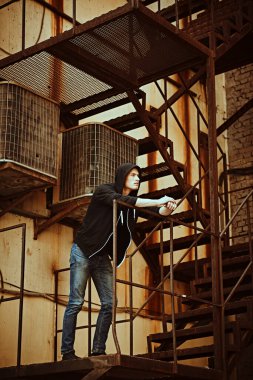 Büyük şehirde hayat. Kapüşonlu ve kotlu yakışıklı genç adam metal endüstriyel bir merdivende duruyor. Kentsel grunge tarzı.