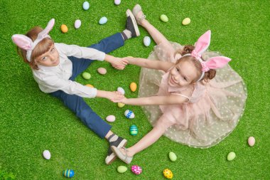 Paskalya tatili. Sevimli gülümseyen çocuk ve tavşan kulaklı bir kız renkli yumurtalarla yeşil bir çimenlikte oturuyorlar.. 