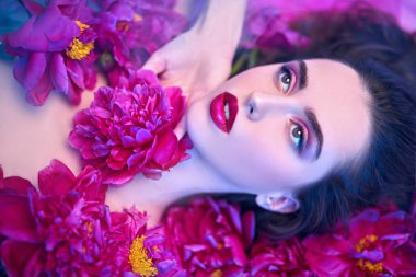 Parfüm ve güzellik ürünleri. Şakayık çiçeklerin arasında uzanan parlak makyajlı güzel esmer bir kadının portresi.. 