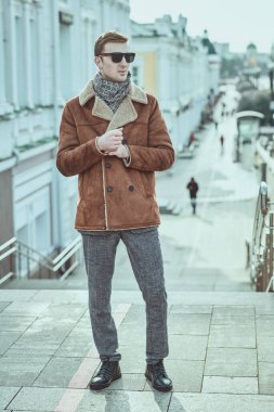 Erkek tarzı. Yakışıklı, takım elbiseli ve kışlık ceketli bir adam şehrin caddesinde duruyor. Kış modası, giysi ve aksesuarlar.