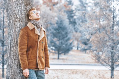 Gözlüklü ve paltolu yakışıklı bir adamın portresi ağaca yaslanmış ve düşünceli bir şekilde gökyüzüne bakıyor..