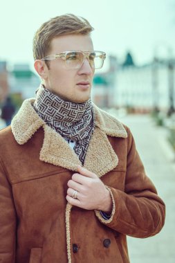 Kışlık ceketli ve atkılı yakışıklı genç adam şehrin sokaklarında duruyor. Kış modası, giysi ve aksesuarlar. Erkek stili. 