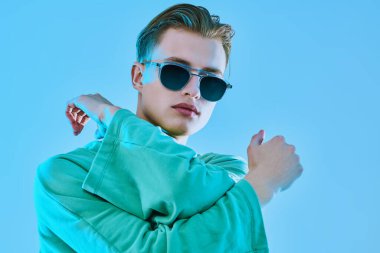 Yakışıklı genç adam modaya uygun kazak ve mavi arka planda güneş gözlüğü ile poz veriyor. Gözlük modası. Stüdyo resmi.