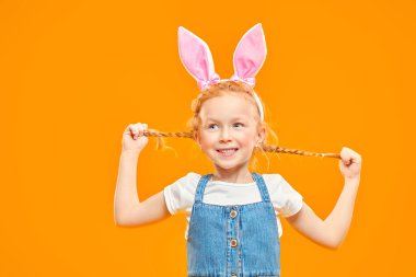 Komik kızıl saçlı kız örgülerini tutuyor ve gülüyor. Mutlu bir çocukluk. Paskalya tatili. Sarı arka planda stüdyo resmi.