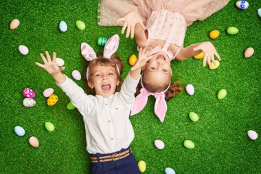 Şirin, gülümseyen bir oğlan ve tavşan kulaklı bir kız renkli yumurtalarla yeşil bir çimenlikte uzanıyorlar. Paskalya tatili.