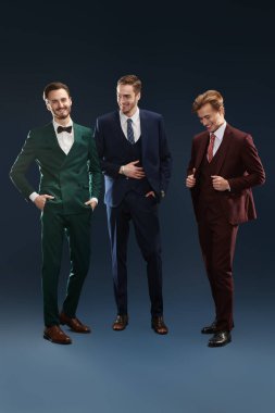 Zarif takım elbiseli üç yakışıklı erkek bir arada durup gülüyor. Üç arkadaş. Erkek modası. Koyu mavi arkaplan.