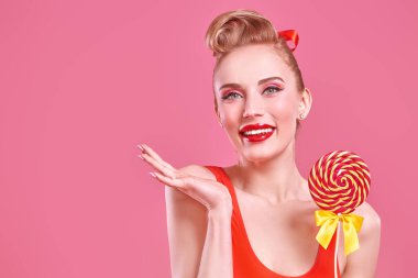 Pin-up güzellik konsepti. Makyajlı güzel sarışın bir kadının portresi. Pembe arka planda lolipopla poz veriyor. Sevgililer Günü. Boşluğu kopyala.