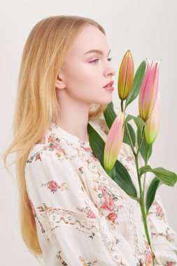 Bahar ve yazın ilham kaynağı. Yazlık bluz giyen güzel sarışın kız beyaz arka planda zambak çiçekleriyle poz veriyor. Pembe renkli açık renk makyaj. Parfüm ve kozmetik.