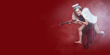 Kırmızı arka planda yaylı tüfek ve okla uçan yakışıklı bir aşk tanrısının tam boy portresi. Aşk Tanrısı. Sevgililer Günü ve Aşk.