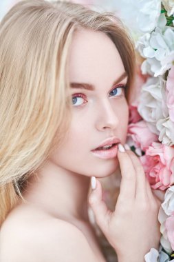 Bahar kızı. Açık renk pembe makyajlı güzel bir genç kadının portresi ve çiçek açan güllerin arka planında sarı saçlar. Güzellik konsepti. 