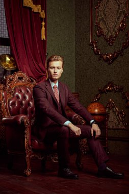Resmi takım elbiseli yakışıklı bir gencin portresi klasik iç mimarisi olan lüks dairelerde bir koltukta oturur. Erkek güzelliği, moda.