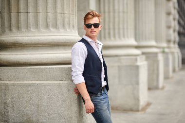 Yakışıklı genç adam eski kasabadaki sütunların arka planında güneş gözlükleriyle yürüyor. Yaz modası. 