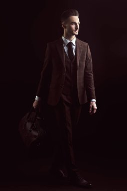 Full length portrait of a handsome man in elegant classic suit and tie posing with leather bag on a black background. Business style. Men's fashion, accessories. clipart