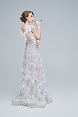 Ağızlıkta sigara içen lüks beyaz elbiseli güzel ve şık bir genç kadının tam boy portresi. Moda fotoğrafı. Gelinlik. Hollywood tarzı.