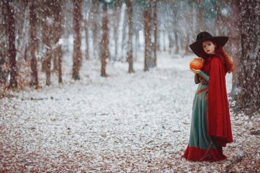 Şapkalı çekici genç cadı karlı ormanın derinliklerinde balkabağı tutuyor. Peri masalları. Cadılar Bayramı. 