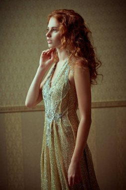 Kaliteli altın duvar kâğıdı arka planda lüks bir elbiseyle poz veren güzel kızıl saçlı çekici bir genç kadının portresi. Güzellik, akşam modası.. 