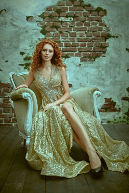 Güzellik, moda konsepti. Güzel kızıl saçlı genç bir kadının portresi. Lüks altın bir elbise içinde, eski bir koltukta poz veriyor.. 