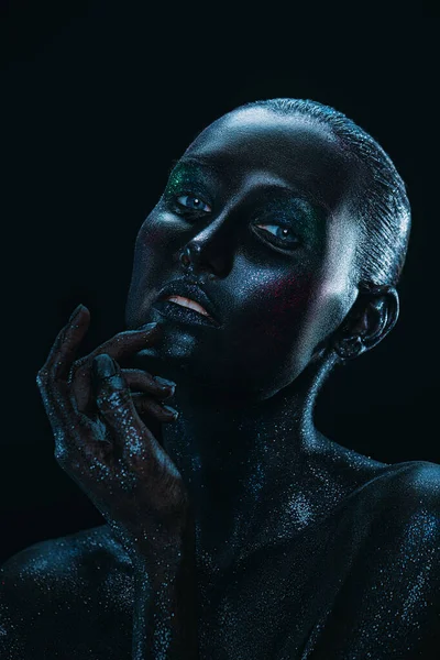 Art beauty portrait. Perfect fashion model girl with black painted skin and sparkling glitter makeup posing in dark with light. Makeup and cosmetics.