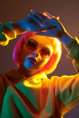 Parlak makyajlı, pembe saçlı, kırmızı güneş gözlüklü, sarı takım elbiseli, karışık renkli ışık saçan modern bir kız. Sportif şıklıkta. Gençlik modası ve güzellik eğilimleri. 