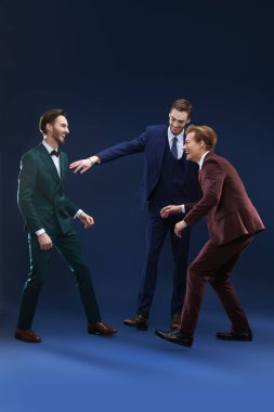 Klasik kostümlü üç genç adam stüdyonun karanlık arka planında durup gülüyor..