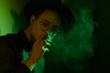 Sigara içen ve etrafı dumanla çevrili yakışıklı bir gencin portresi. Stüdyo portresi koyu yeşil ışıkta.