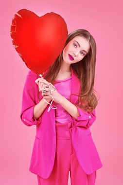 Pembe elbiseli, elinde kalp şeklinde bir balon tutan hoş bir kız. Pembe arka planda stüdyo resmi. 