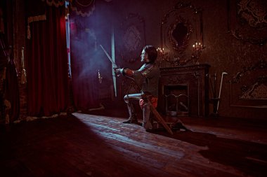 Zırhlı asil şövalye diz çöküp kılıcını kaldırdı ve karanlık kaledeki ışığa baktı. Romantizm. Tam boy portre.