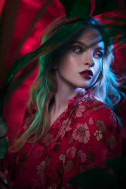 Uzun dalgalı sarı saçlı, parlak makyajlı, tropik yeşillikler arasında kırmızı elbiseli güzel bir manken. Güzellik, moda. Orman fotoshoot. 