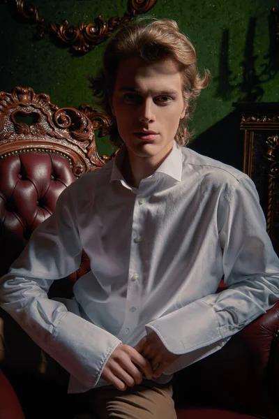 Luxury lifestyle. Portrait of a handsome young man in elegant white shirt sitting in an armchair in a luxury apartments. Men's beauty, fashion.