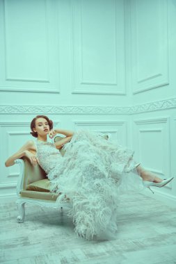 Lüks beyaz bir elbise içinde klasik koltukta poz veren güzel, şık bir kadının portresi. 20 'lerin akşam makyajı ve saç stili.. 