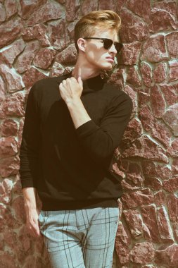 Siyah kazak, kot pantolon ve güneş gözlükleriyle taş duvarın yanında poz veren modaya uygun genç adam modeli. Erkek stili, güzellik. Optikler.