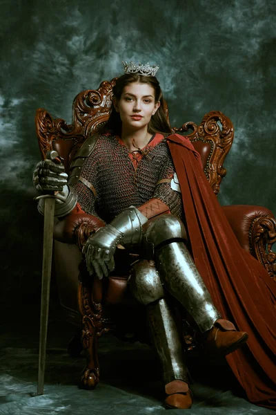 Full length portrait of a medieval queen in knightly armor sitting on a throne on a grunge background. History of the Middle Ages.