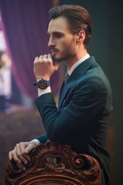 Zarif klasik takım elbiseli yakışıklı genç adam lüks bir dairede eski bir koltuğun yanında poz veriyor. Erkek güzelliği, moda. Lüks yaşam tarzı. 