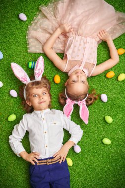 Şirin, gülümseyen bir oğlan ve tavşan kulaklı bir kız renkli yumurtalarla yeşil bir çimenlikte uzanıyorlar. Paskalya tatili.