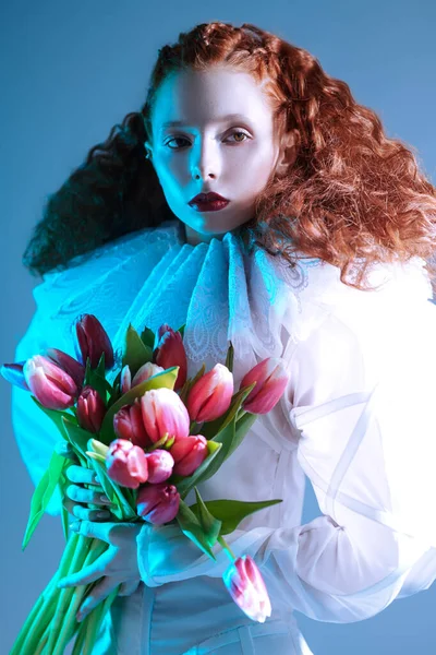 Portrait of a refined fashion model girl with long red curly hair posing in a white haute couture dress with late renaissance ruffled collar and tulips. Art fashion history.