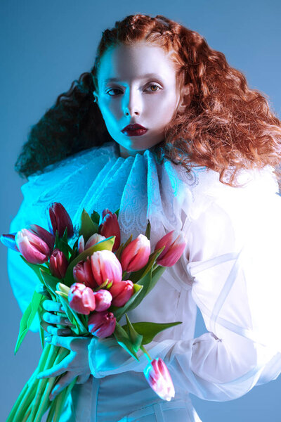 Portrait of a refined fashion model girl with long red curly hair posing in a white haute couture dress with late renaissance ruffled collar and tulips. Art fashion history. 