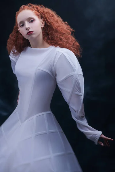 High fashion art. Sophisticated female model with lush red curly hair posing in a white art dress. A studio portrait on a vintage background.