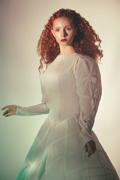 Art portrait of a refined fashion model girl with lush red curly hair posing in a white haute couture dress. Studio shot. 