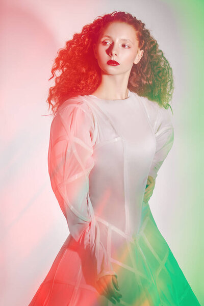 Portrait of a refined fashion model girl with lush red curly hair posing in a white haute couture dress. Art and fashion. Studio shot in mixed color light. 
