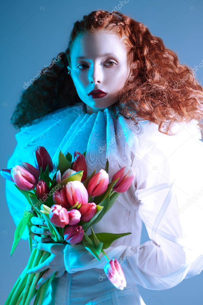Portrait of a refined fashion model girl with long red curly hair posing in a white haute couture dress with late renaissance ruffled collar and tulips. Art fashion history. 