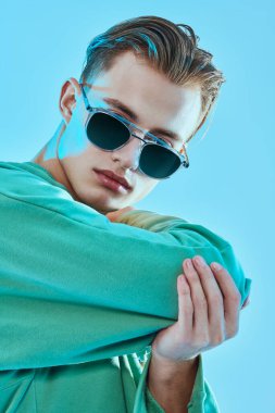 Yakışıklı genç adam modaya uygun kazak ve mavi arka planda güneş gözlüğü ile poz veriyor. Gözlük modası. Stüdyo resmi.