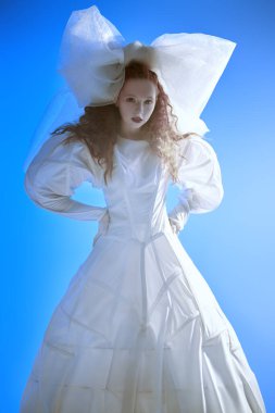 Fashion art portrait of a girl looking like a doll with lush red curly hair posing in a white haute couture dress and a huge bow. Studio portrait on blue background.