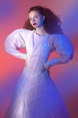 Sophisticated female model with lush red curly hair posing in a white art dress. A studio portrait with mixed red color light. Fashion art. 
