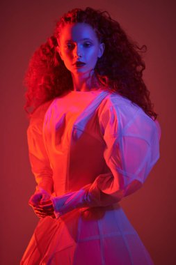 Portrait of a sophisticated female model with lush red curly hair posing in a white art dress. A studio portrait with mixed red color light. Fashion art. 