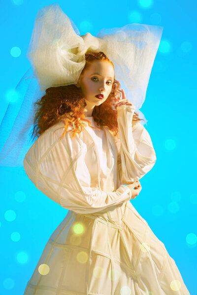 Fashion art portrait of a girl looking like a doll with lush red curly hair posing in a white haute couture dress and a huge bow. Studio portrait.