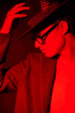 Attractive sexual young man posing in a coat on a naked torso, a hat and fashionable glasses. Fashion portrait in dark red light. Optics for men.