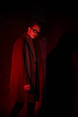 Portrait of a handsome sexual young man posing in a coat and fashionable glasses in dark red light. Men's fashion and optics.