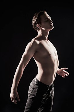 Handsome athletic man with beautiful muscular body poses shirtless in the dark, turning and aiming for the light. Black background. Men's beauty and health.