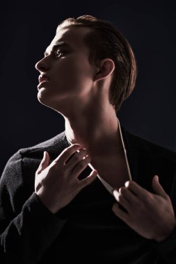 Portrait of emotional young man pulling back the collar of his clothes at studio in dark lighting. Men's beauty. Black background. 